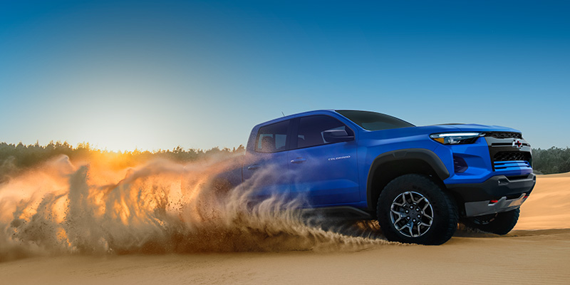 A blue 2026 Chevrolet Colorado pickup truck drives through sandy terrain, kicking up a large cloud of dust as it speeds across a desert landscape under a clear sky.