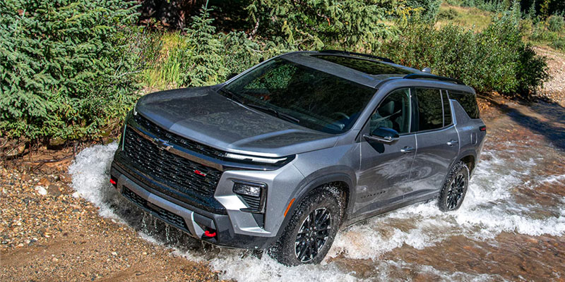 A gray 2026 Chevrolet Traverse SUV drives through a shallow rocky stream, with water splashing around the tires and trees lining the background.