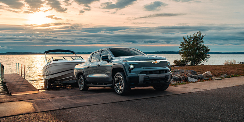 A 2026 Chevrolet Silverado EV pickup truck is parked at a lakeside boat ramp at sunset, with a boat on a trailer behind it and calm water stretching into the distance under a cloudy sky.